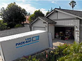 A 1-800-PACK-RAT portable storage container sits open in the driveway of a suburban home with its garage door open on a bright sunny day