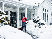 A man shovels snow off his front porch and sidewalk leading to the front of his home