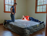 A woman rests on a mattress while her husband stands next to the mattress with a cover on it in an empty room after moving it in