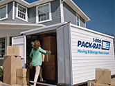 A woman packs her 16-foot 1-800-PACK-RAT portable storage container in her home's driveway as boxes fill the container and sit outside it on a bright, sunny day.