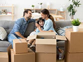 A mother and father sit with their daughter on their couch smiling and laughing as they unpack boxes inside the living room of their new home