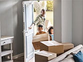 A man opens the door for his two children carrying boxes into the new home with a view of unpacked boxes sitting near the doorway