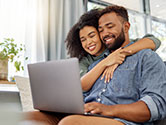 A couple sits on their couch in their home's living room smiling while working on their laptop