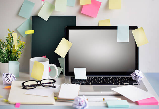 A cluttered work desk where a laptop with sticky notes on it sits