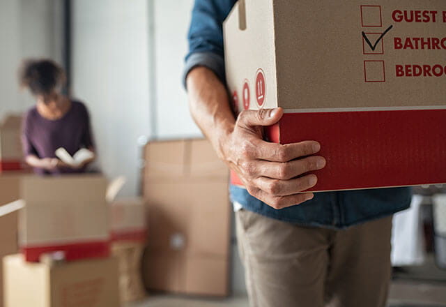 A man carries a moving box while a woman in the background packs boxes