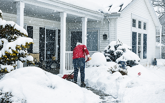 A man shovels snow off his front porch and sidewalk leading to the front of his home