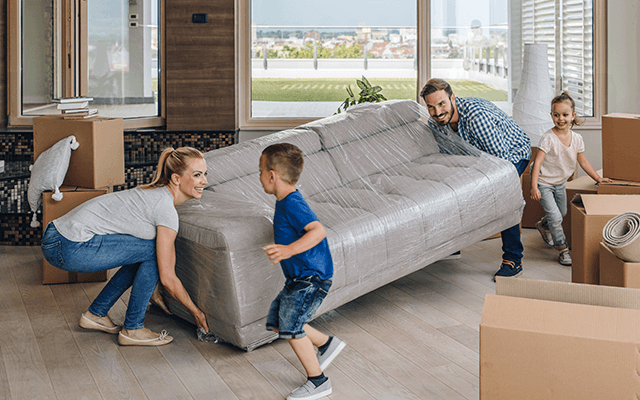 A man and woman lift a couch to move while their children run around the room