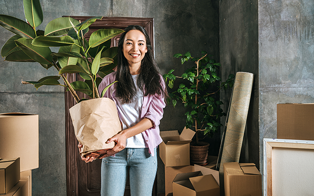 A woman smiles as she holds her houseplant in her hands surrounded by packed and open boxes inside her home