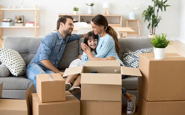 A mother and father sit with their daughter on their couch smiling and laughing as they unpack boxes inside the living room of their new home