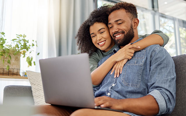 A couple sits on their couch in their home's living room smiling while working on their laptop