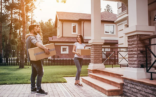 A man holding his child and a moving box stands near his wife on the sidewalk of their new home in a nice neighborhood at sunset