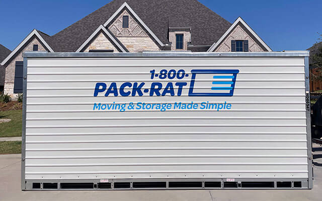 Side view of a 16-foot 1-800-PACK-RAT portable storage container that is being delivered to the street-front of a suburban home on a bright sunny day