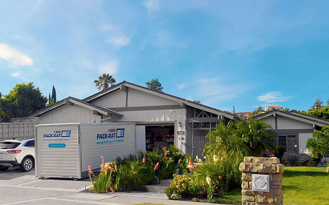 A 1-800-PACK-RAT portable storage container sits open in front of an open garage with a white SUV parked next to it in the driveway of a suburban home on a bright sunny day