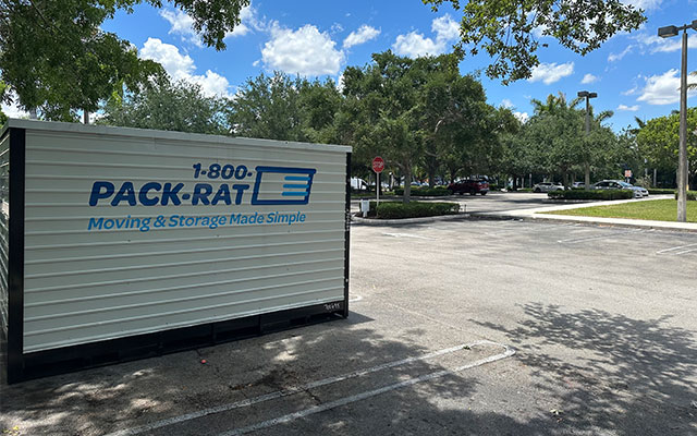 A side view of a 1-800-PACK-RAT portable storage container sitting in the parking lot of a business on a bright sunny day