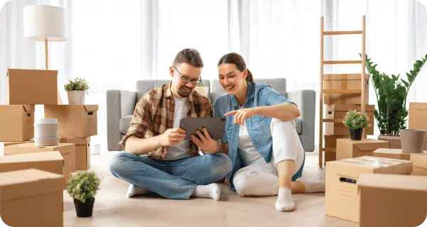 A couple sits on a floor looking at a tablet computer while being surrounded by moving boxes and some indoor plants