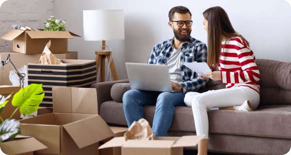 A happy couple sits on their couch discussing something while surrounded by half-packed boxes