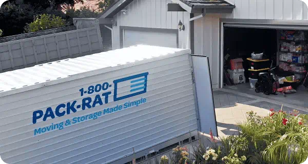 A 1-800-PACK-RAT portable storage container sits open in the driveway of a home with the garage open