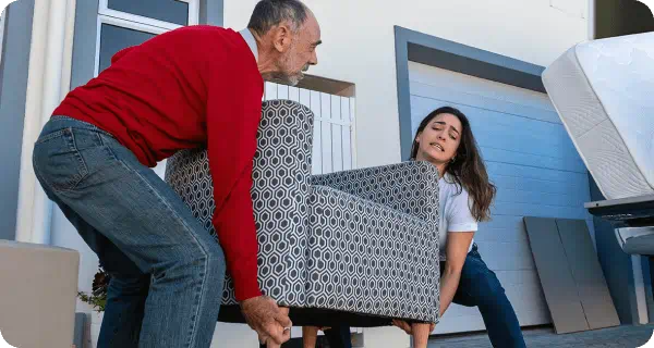 A man and a woman struggle to carry a chair to their moving truck while other moving items lay around them