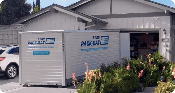 A 1-800-PACK-RAT portable storage container sits open in the driveway of a suburban home with it's garage door open