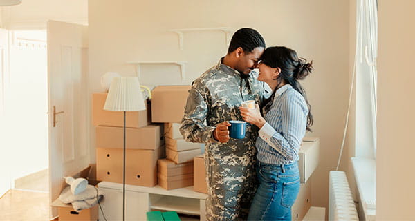 A military member and his spouse hug each other in their new home with unpacked moving boxes sitting behind them