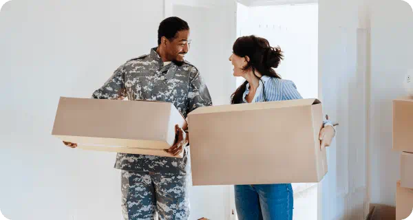 A military service member and his wife smile and laugh as they carry boxes into their new home