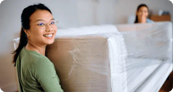 Two women are carrying a shrink-wrapped couch out of a home