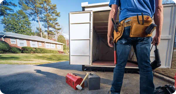 A contractor with his toolbelt stands in front of an open portable storage container sitting on a neighborhood road in front of a house