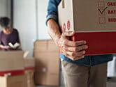 A man carries a moving box while a woman in the background packs boxes