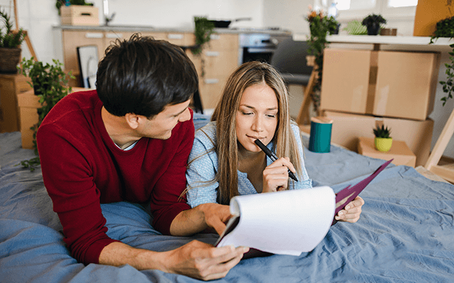 A happy couple makes a list on a notepad surrounded in their room by packed boxes