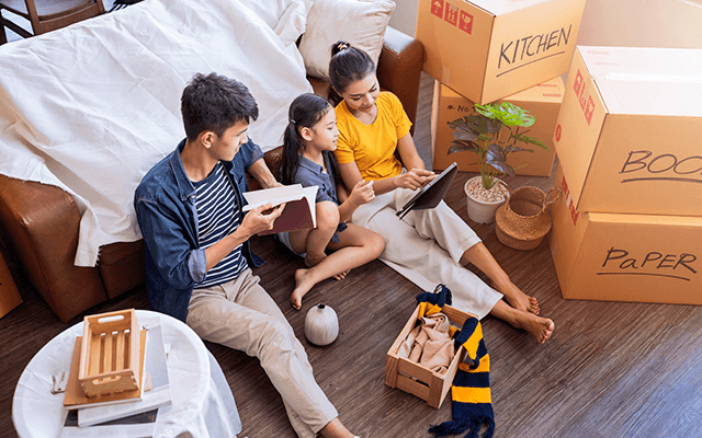 A family sits on the floor of their new home looking at a tablet surrounded by marked packed boxes