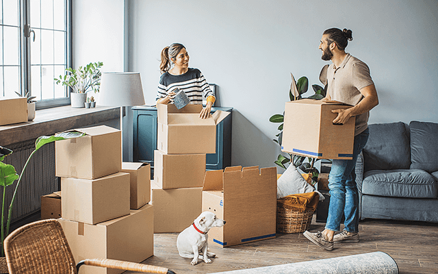 A couple unpacks moving boxes in their new home while their dog sits on the floor