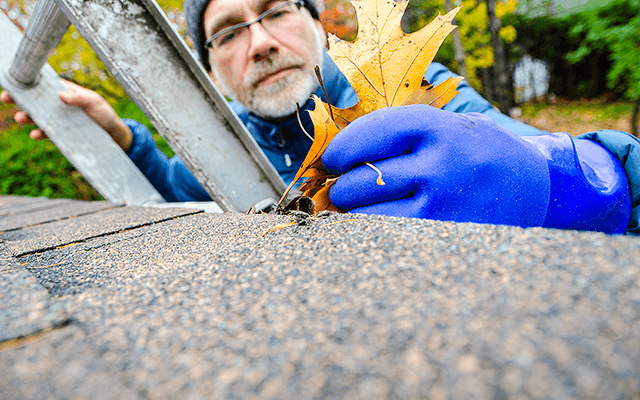 A homeowner cleans leaves out of his home's gutters while standing on a ladder on a Fall day