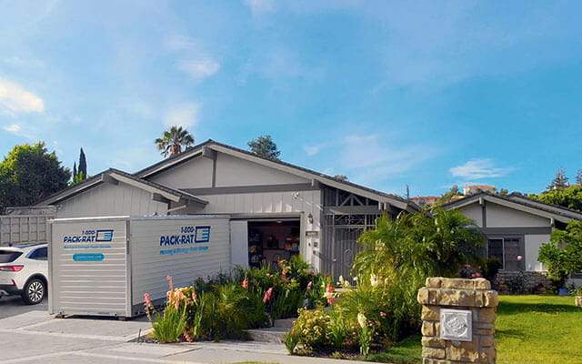 A 1-800-PACK-RAT portable storage container sits open in front of a home's open garage in the driveway on a bright sunny day