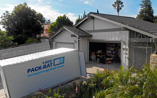 A 1-800-PACK-RAT portable storage container sits open in the driveway of a suburban home with its garage door open on a bright sunny day