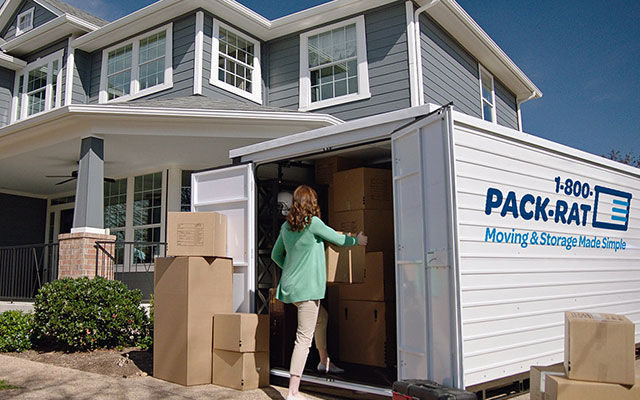 A woman packs her 16-foot 1-800-PACK-RAT portable storage container in her home's driveway as boxes fill the container and sit outside it on a bright, sunny day.