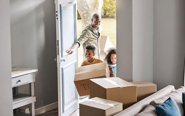 A man opens the door for his two children carrying boxes into the new home with a view of unpacked boxes sitting near the doorway