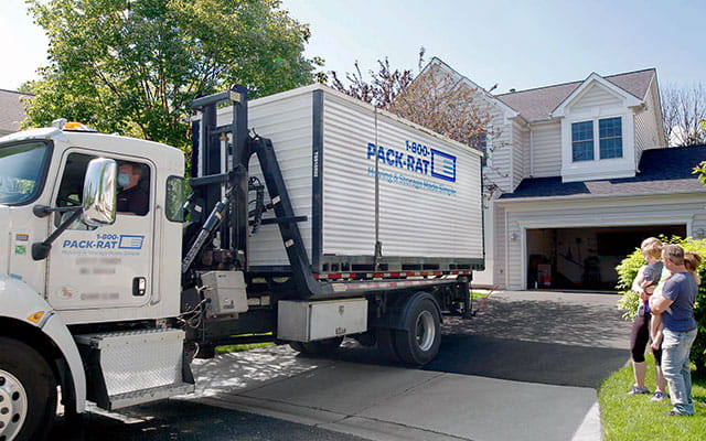 View of a 1-800-PACK-RAT driver backing in a mini-mover truck with a portable storage container on the back of it into the driveway of a customer as the customers watch on a bright sunny day