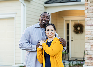 A middle-aged couple stands happily in front of their home