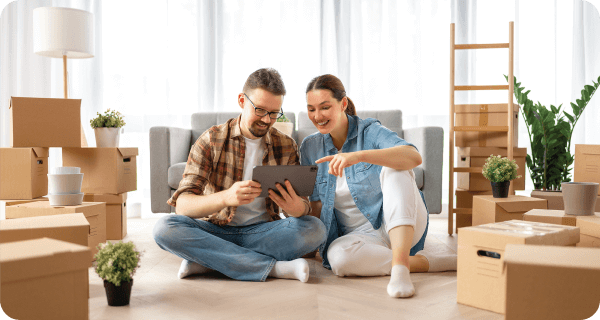 A couple sits on a floor looking at a tablet computer while being surrounded by moving boxes and some indoor plants