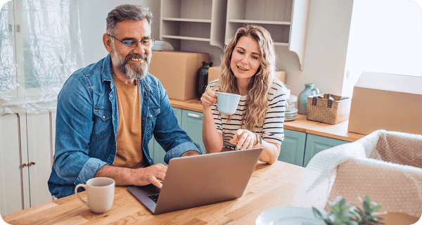 A middle-aged couple sits at their table surrounded by packed boxes while looking at something on their laptop