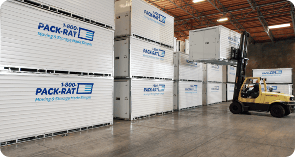 A number of 1-800-PACK-RAT portable storage containers sit in a warehouse while a forklift places a container