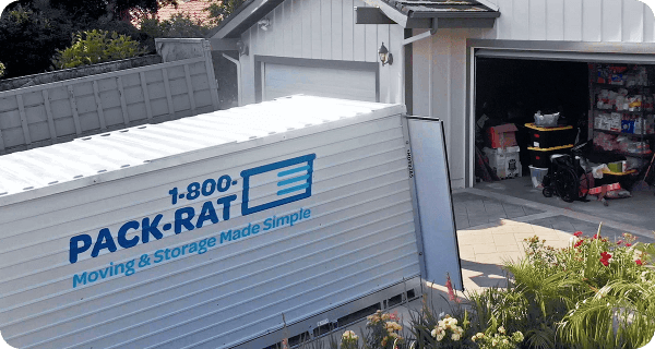 A 1-800-PACK-RAT portable storage container sits open in the driveway of a home with the garage open