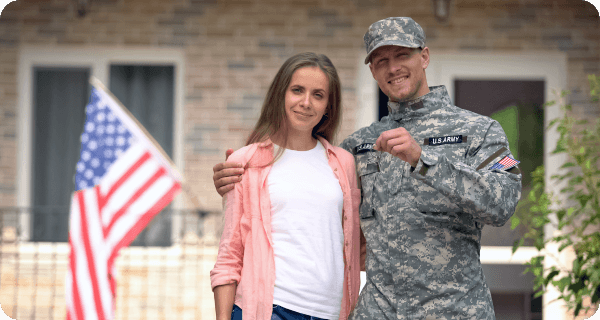 A military couple looks on happily holding the keys to their new home that lays in the background with an American flag present