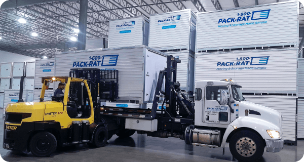 A 1-800-PACK-RAT portable storage container is carefully placed on the back of a mini-mover truck by a forklift inside of a 1-800-PACK-RAT storage facility with many containers in the background