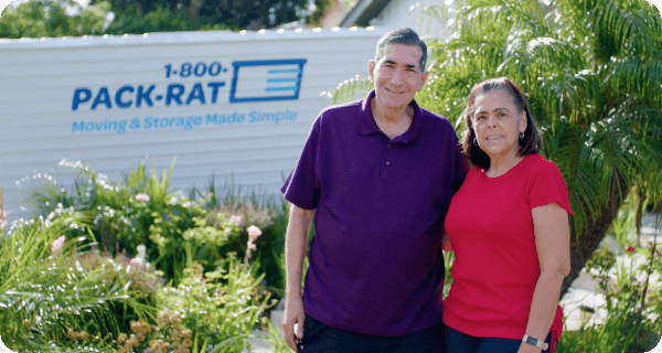 A happy couple stands in front of their home with a 1-800-PACK-RAT container sitting in the background behind them