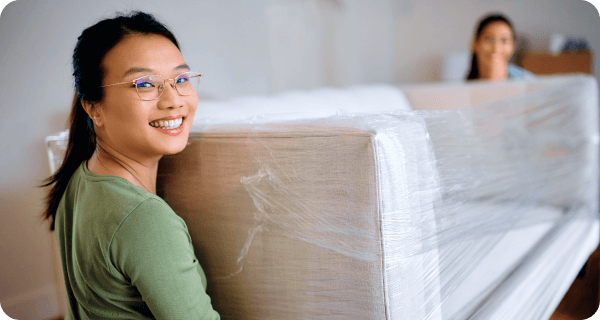 Two women are carrying a shrink-wrapped couch out of a home