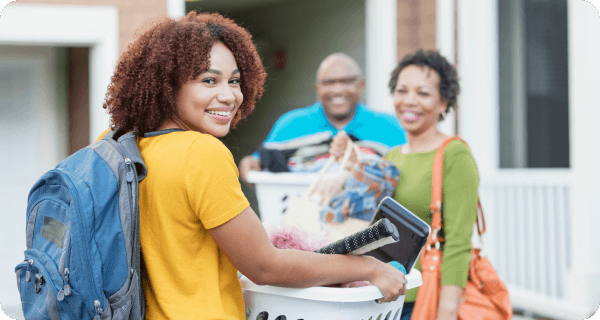 A college student carries a hamper filled with her belongings while her parents are in the background helping her move into her dorm