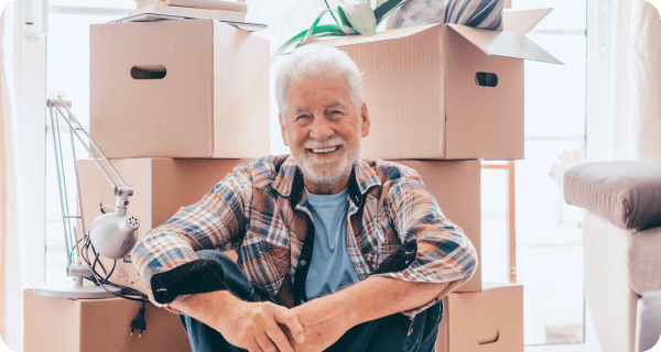 An older man smiles and sits in front of a number of packed boxes on the floor