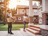 A man holding his child and a moving box stands near his wife on the sidewalk of their new home in a nice neighborhood at sunset