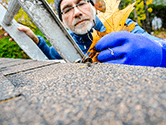 A homeowner cleans leaves out of his home's gutters while standing on a ladder on a Fall day